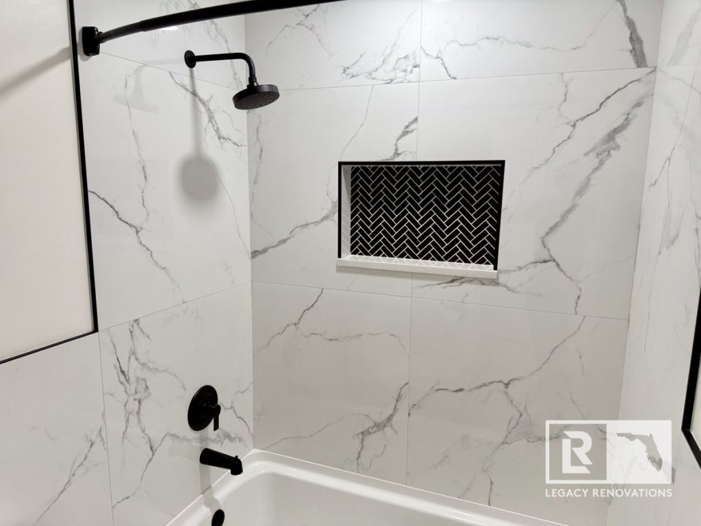 Remodeled bathtub and shower featuring large-format marble-look porcelain tile, matte black fixtures, and a recessed herringbone shower niche.