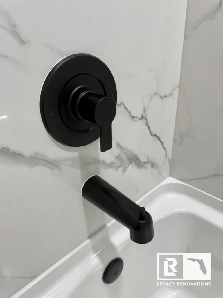 Close-up of a matte black bathtub faucet, tub spout, and shower valve installed on large-format marble-look wall tile.