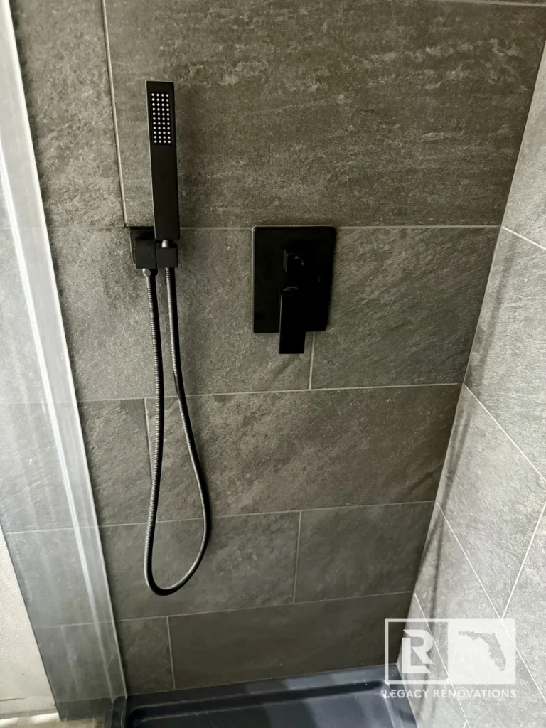 Matte black shower valve and handheld shower sprayer installed on large-format gray porcelain tile in a remodeled bathroom.