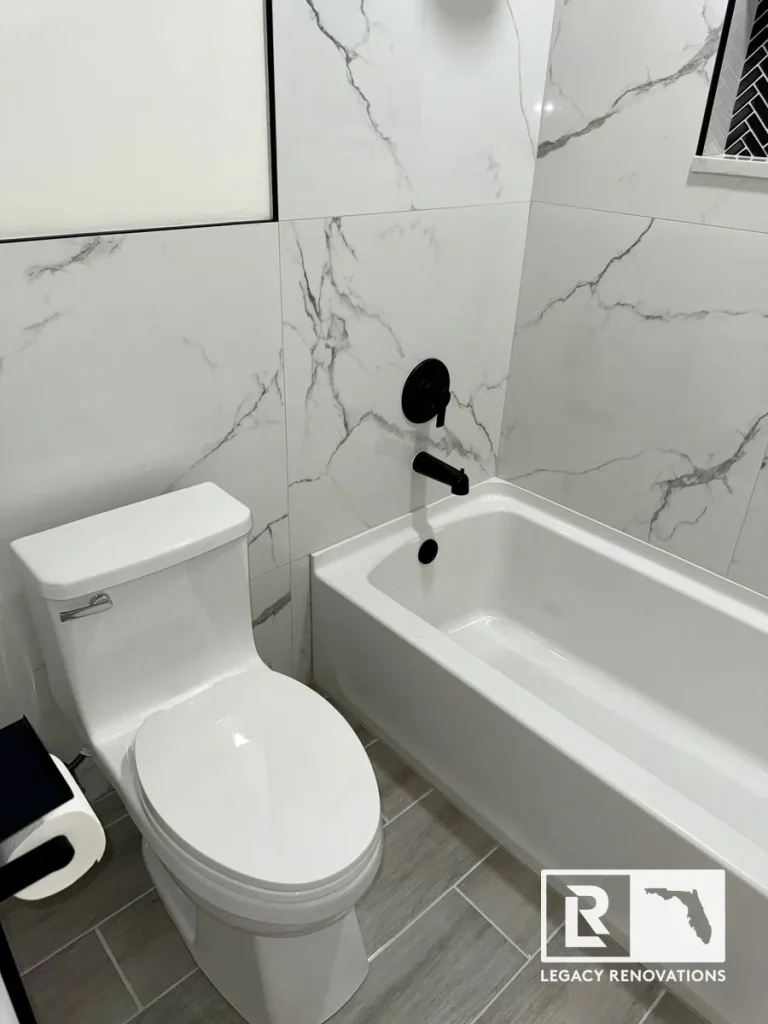 Modern bathroom featuring a white bathtub with a marble-look tile surround, matte black tub faucet, and contemporary gray floor tile.