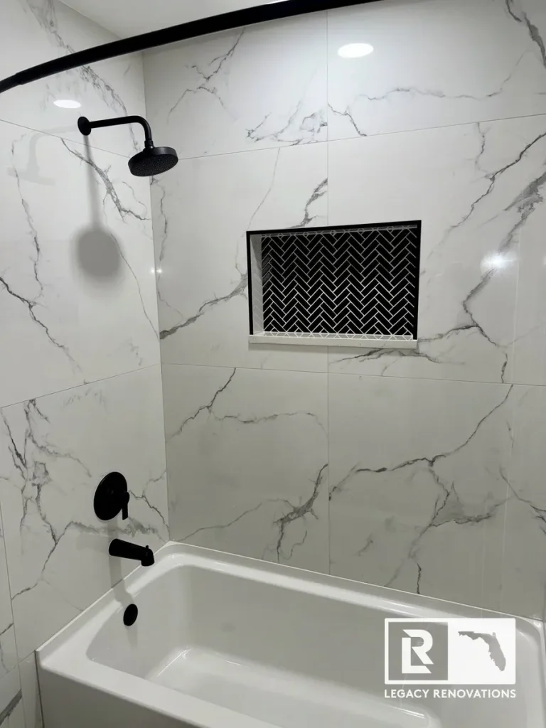 Modern bathtub and shower remodel with large-format marble-look tile, matte black fixtures, and a recessed herringbone mosaic shower niche.