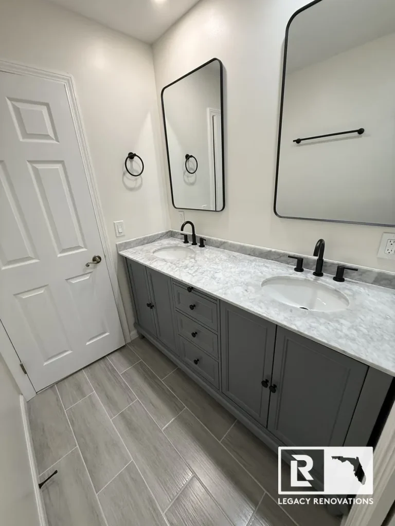 Modern bathroom remodel featuring a gray double vanity, marble-look countertop, black fixtures, framed mirrors, and gray wood-look tile flooring.
