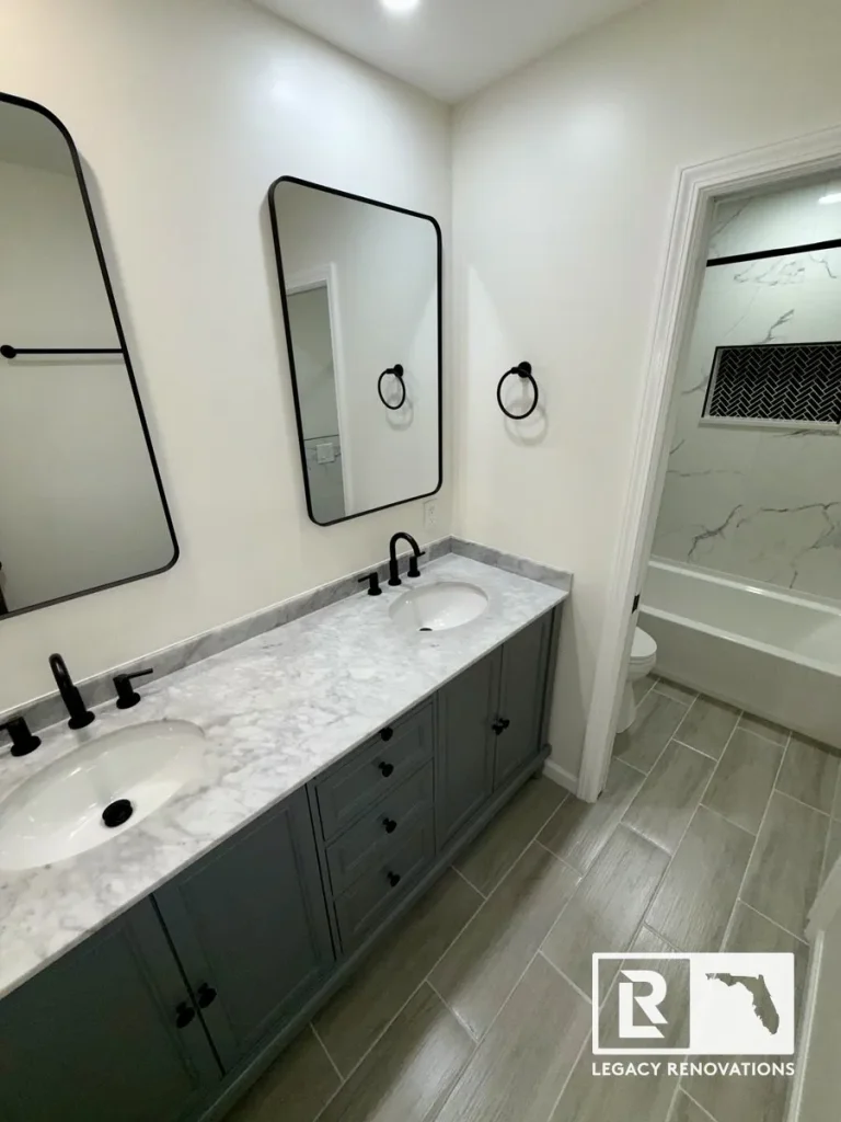 Modern bathroom remodel featuring a gray double vanity, marble-look countertop, black faucets, framed mirrors, and an adjoining bathtub with marble-look tile.