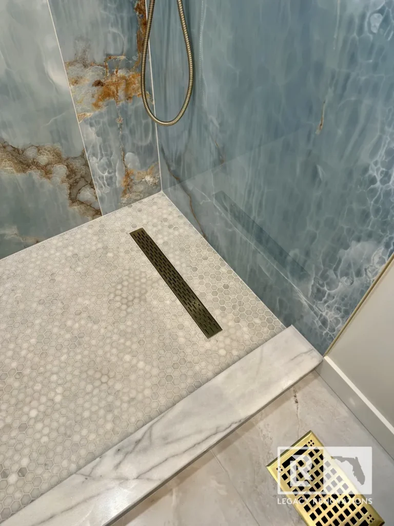 Walk-in shower featuring light blue onyx marble wall panels, white hexagon mosaic floor tiles, a brushed brass linear drain, and a white marble curb.