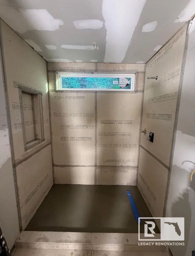 Walk-in shower remodel in progress showing cement backer board walls, shower niche cutout, horizontal transom window, and pre-pitched mortar pan.