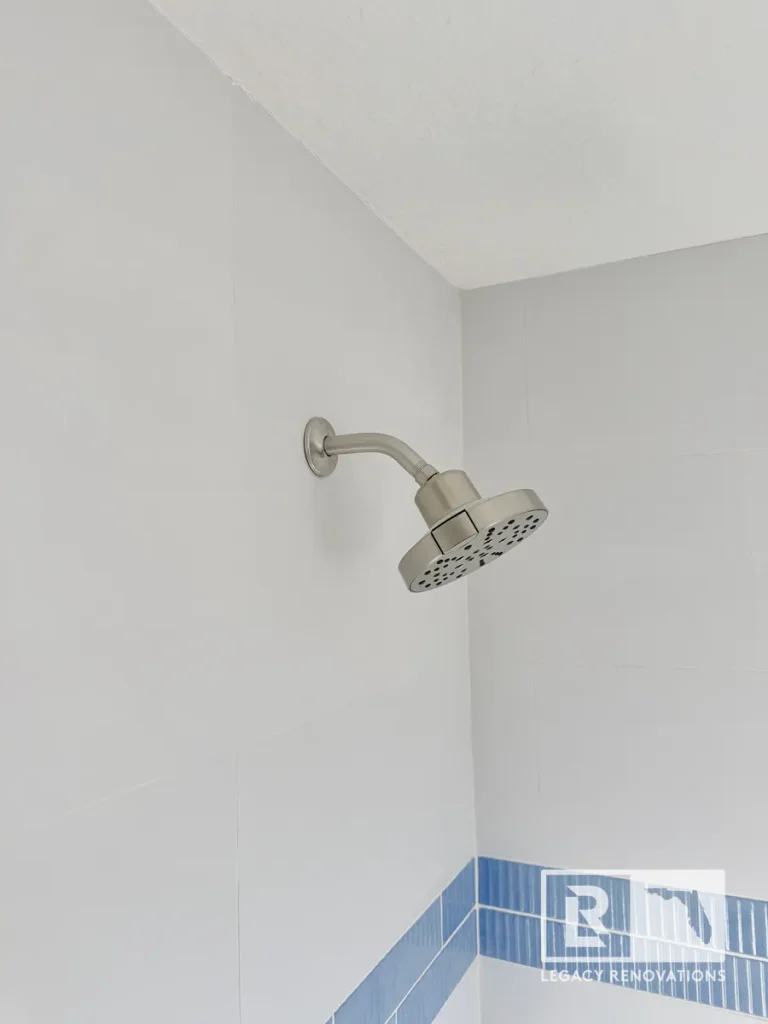 Brushed nickel showerhead mounted on large white tile walls with a blue mosaic tile accent strip in a Jacksonville, FL bathroom renovation.