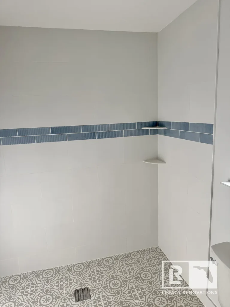 Completed shower corner in Jacksonville, FL featuring white large-format wall tile, blue mosaic accent band, two corner shelves, and patterned floor tile.