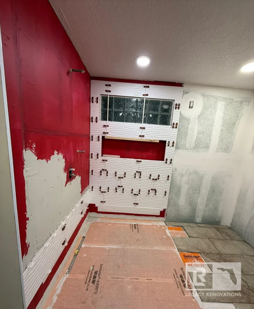 Walk-in shower under construction with textured white wall tile, RedGard waterproofing, Schluter waterproofing membrane, recessed niche, and glass block window during a bathroom remodel in Jacksonville, FL.