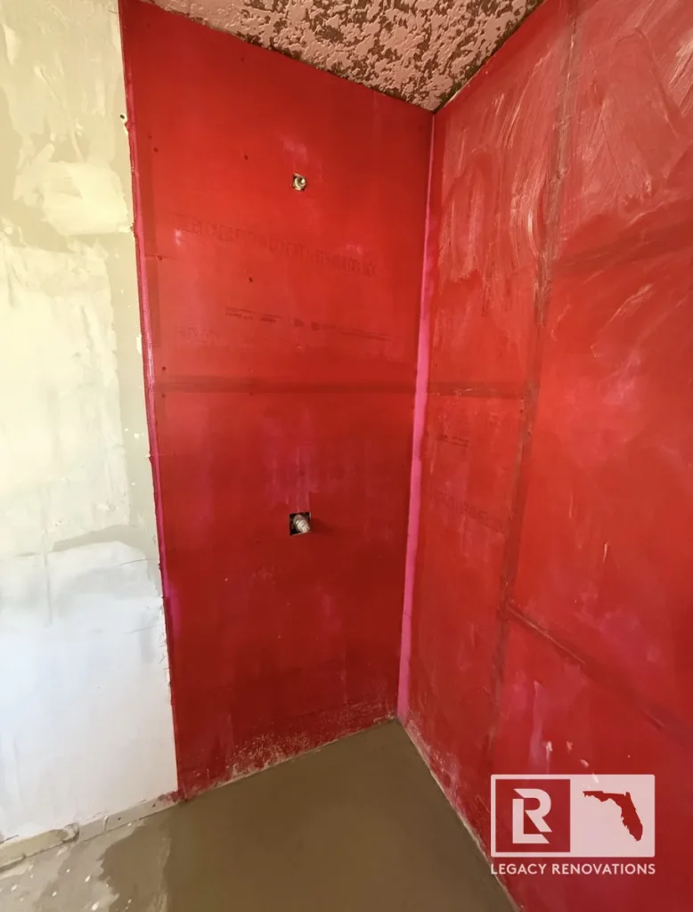 In-progress shower stall preparation in Jacksonville, FL showing red liquid waterproofing membrane applied to backer board walls with a sloped mortar floor pan.