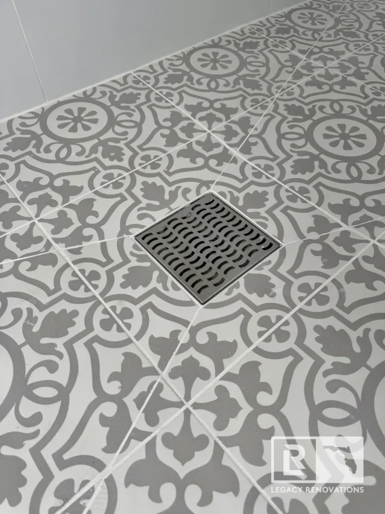 Square brushed stainless steel shower drain installed in a grey and white patterned mosaic tile shower floor in Jacksonville, FL.