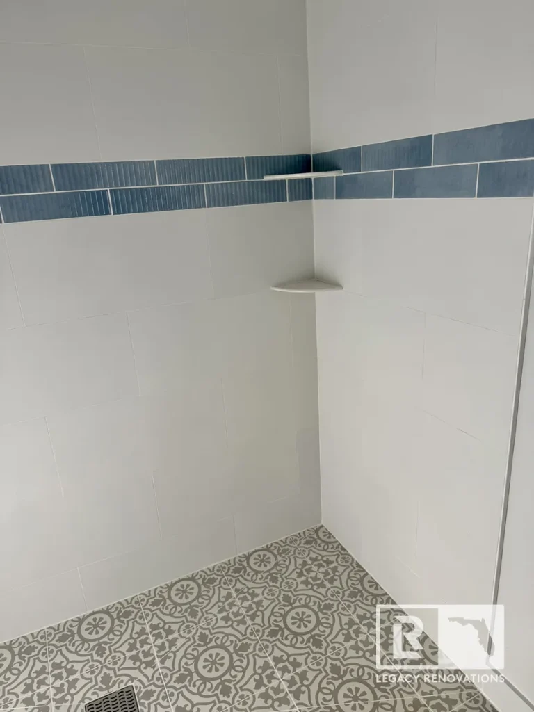 Corner of a walk-in tile shower in Jacksonville, FL featuring white wall tile, blue mosaic accent strip, floating corner shelves, and patterned floor tile.