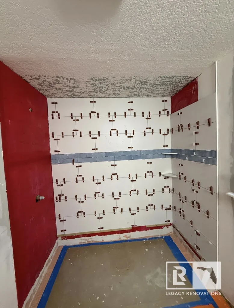 In-progress shower wall tile installation in Jacksonville, FL showing tile leveling spacers, blue accent mosaic band, and red waterproofing membrane.