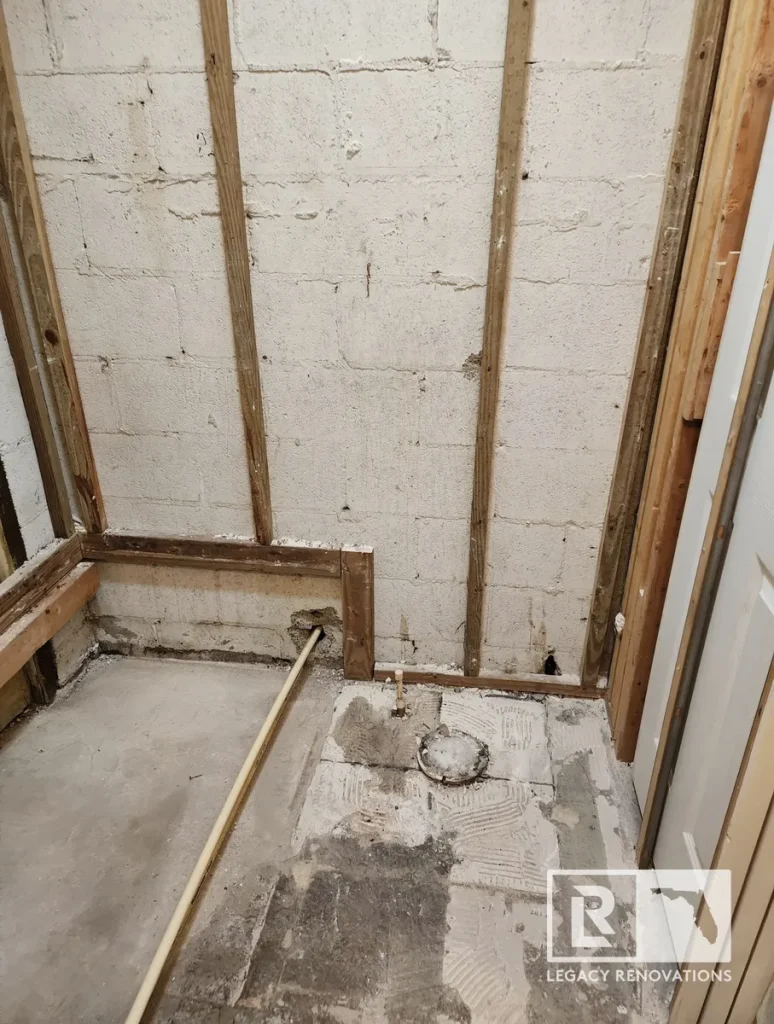A bathroom during the early gut renovation phase, showing exposed concrete block walls, wood studs, a bare concrete floor, and floor drain lines by Legacy Renovations in Jacksonville, FL.