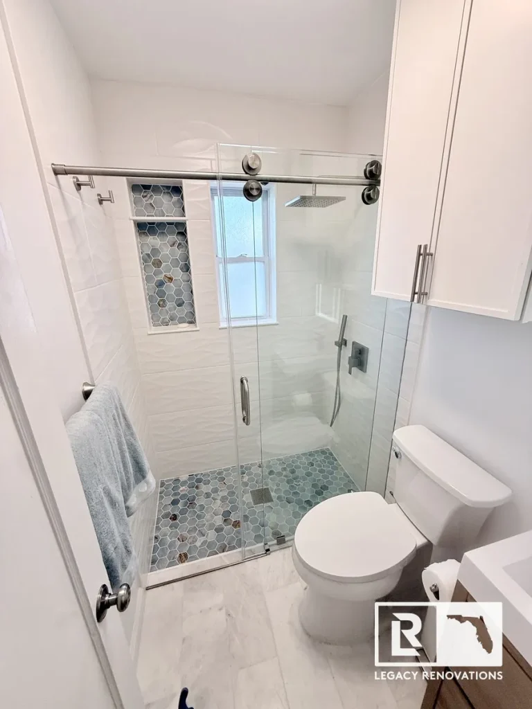 A fully renovated small bathroom showcasing a walk-in shower with sliding glass doors, white wavy wall tile, blue hexagon floor tile, a white toilet, and a matching storage cabinet by Legacy Renovations in Jacksonville, FL.