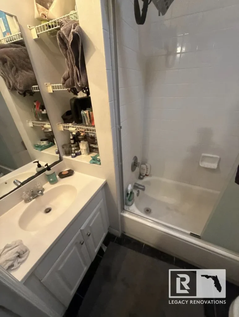 Original bathroom with a white vanity, built-in storage shelves, and bathtub with sliding glass doors before remodeling in Jacksonville, FL.