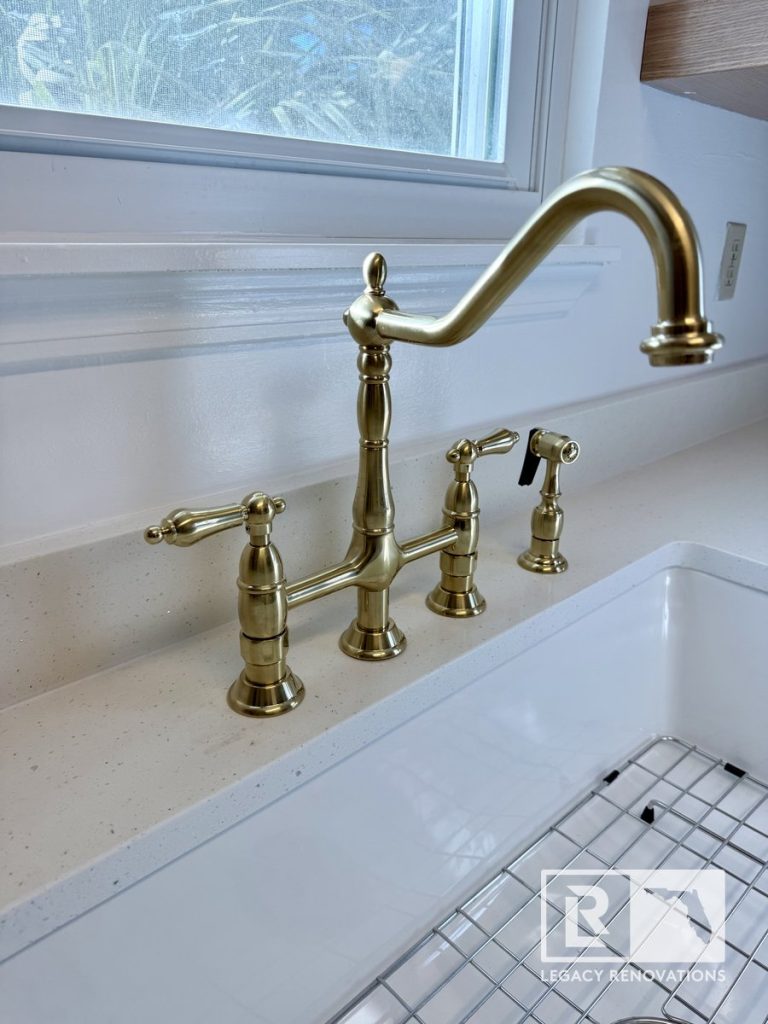 Close-up of an antique brass bridge faucet with dual lever handles and side sprayer installed on a white quartz countertop above a porcelain farmhouse sink.