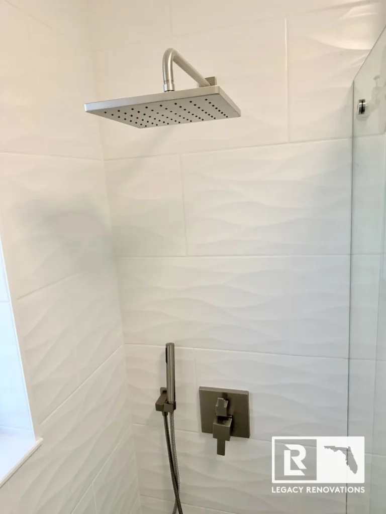 A finished walk-in shower featuring a rectangular brushed nickel rain shower head, a matching handheld wand, and white textured wave-patterned wall tiles by Legacy Renovations in Jacksonville, FL.