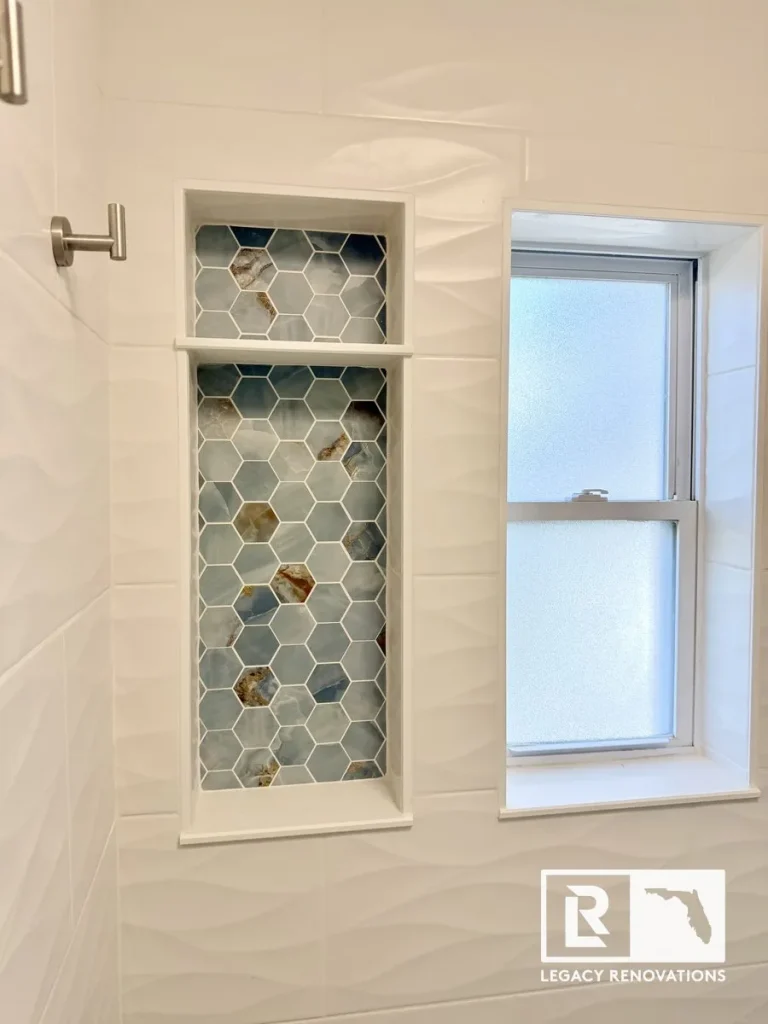 A finished walk-in shower showing a frosted glass window next to a tall recessed niche with two shelves, backed with blue onyx-style hexagon tiles and surrounded by wavy white wall tile, by Legacy Renovations in Jacksonville, FL.