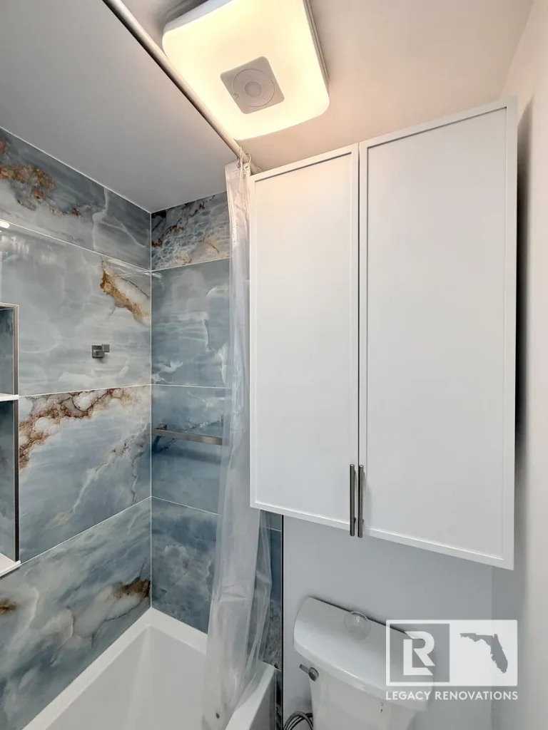 Custom white bathroom storage cabinets above toilet with blue porcelain tile tub surround, recessed shower niche, and modern bathroom remodel in Jacksonville, FL.