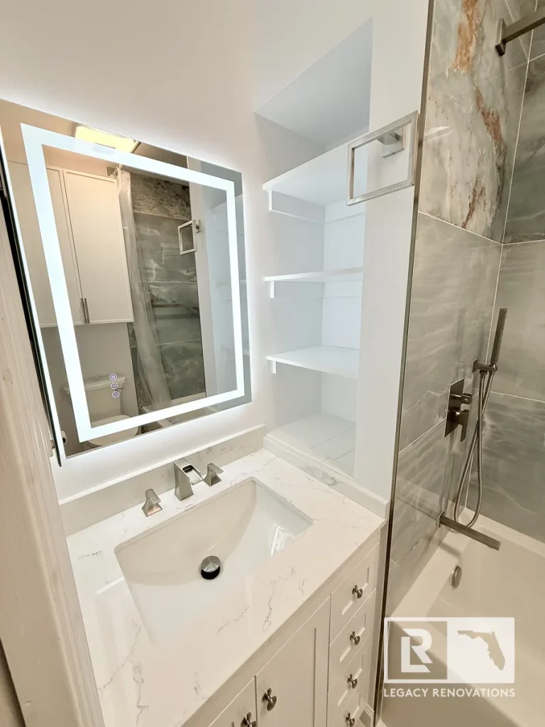 Modern white bathroom vanity with quartz countertop, LED lighted mirror, built-in shelves, frameless glass tub shower, and porcelain tile during a bathroom remodel in Jacksonville, FL.