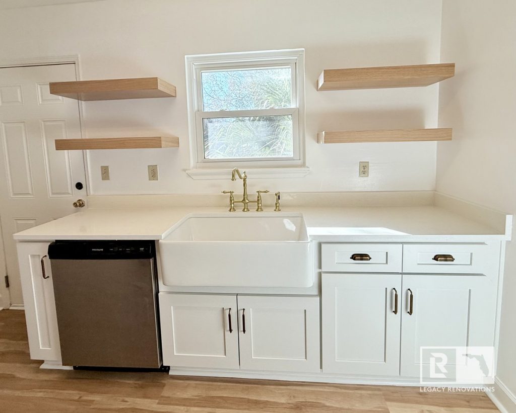 Custom laundry room with white shaker cabinets, farmhouse sink, quartz countertop, floating wood shelves, brushed brass faucet, and stainless steel dishwasher in Jacksonville, FL.