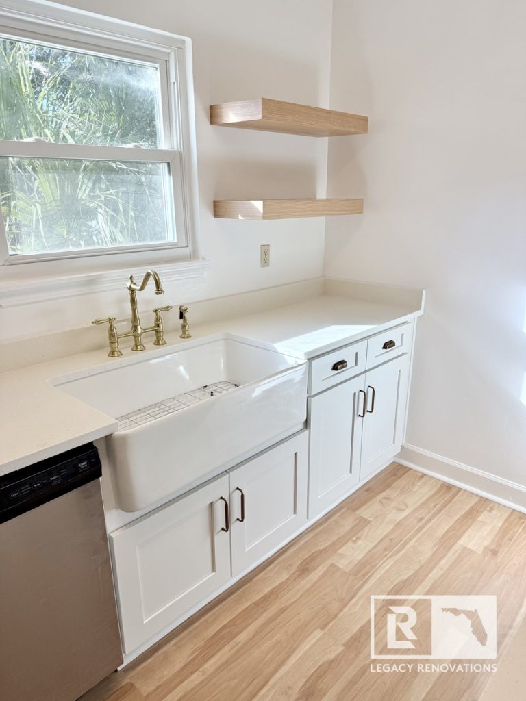 Bright kitchen renovation in Jacksonville, FL featuring a fireclay ceramic sink, antique brass bridge faucet, white recessed-panel cupboards, and open oak floating shelves.