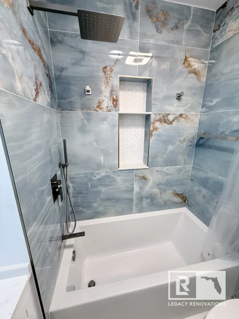 Luxury bathtub and shower remodel with blue porcelain wall tile, recessed shower niche, matte black rainfall showerhead, frameless glass panel, and modern fixtures in Jacksonville, FL.