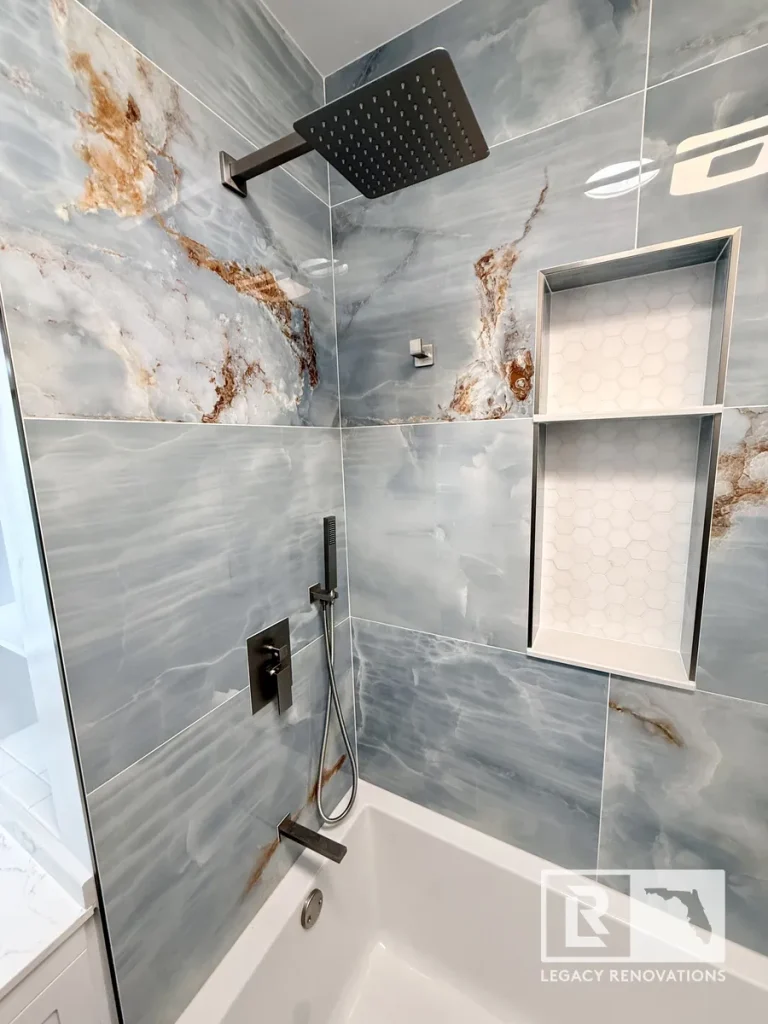 Modern bathtub and shower remodel with large-format marble-look porcelain tile, matte black rainfall shower system, frameless glass panel, and double recessed niche in Jacksonville, FL.