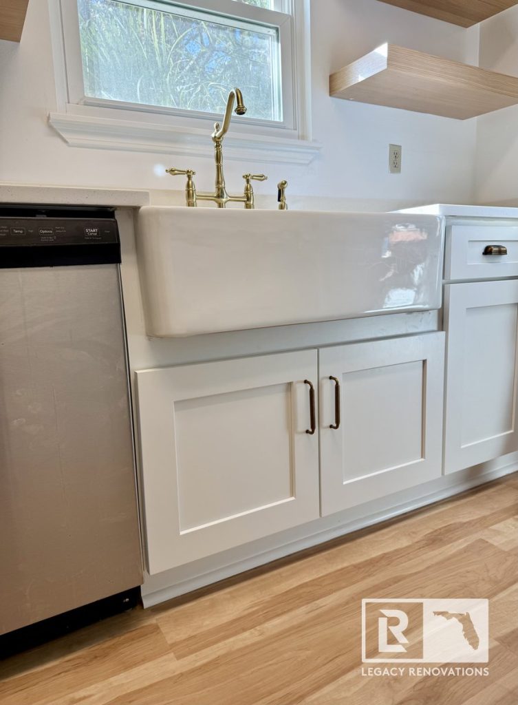 White apron-front farmhouse sink with a vintage brass gooseneck faucet set in custom white shaker cabinets with light wood floors.