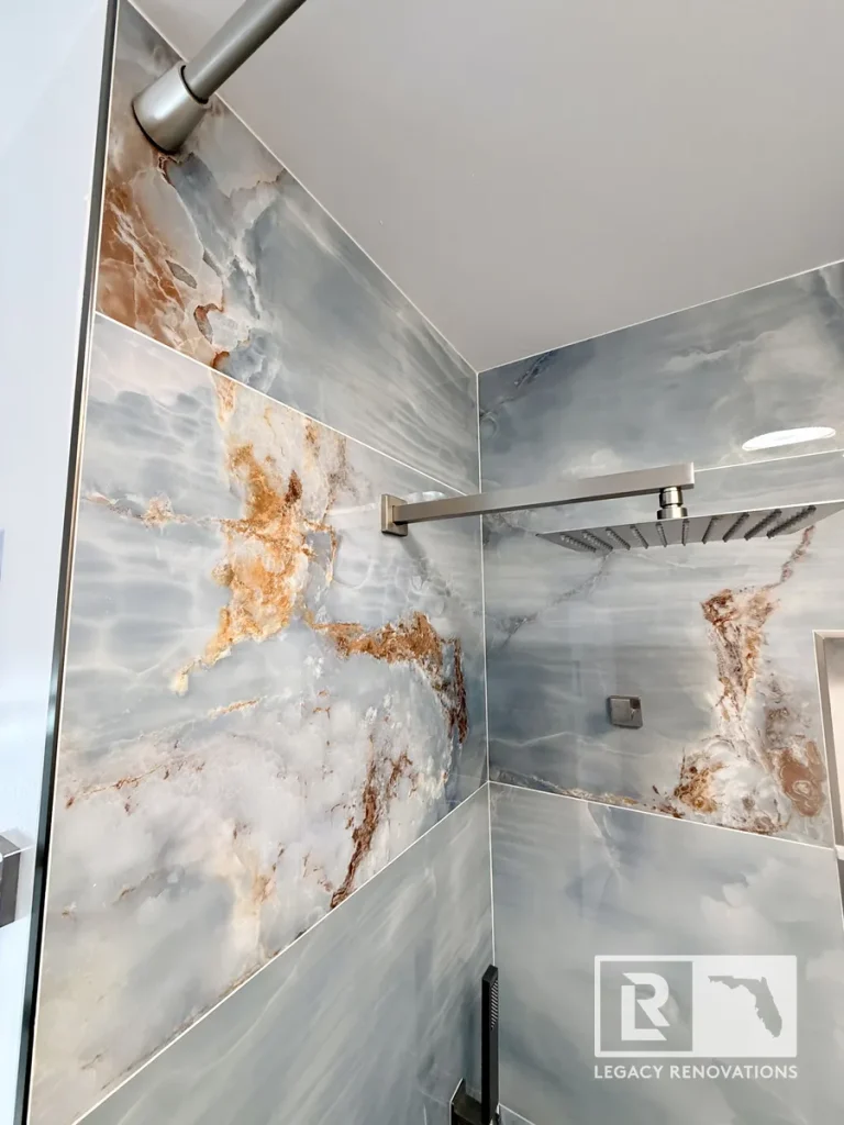 Luxury rainfall shower with blue porcelain marble-look tile, brushed nickel fixtures, frameless glass enclosure, and recessed niche in a custom bathroom remodel in Jacksonville, FL.