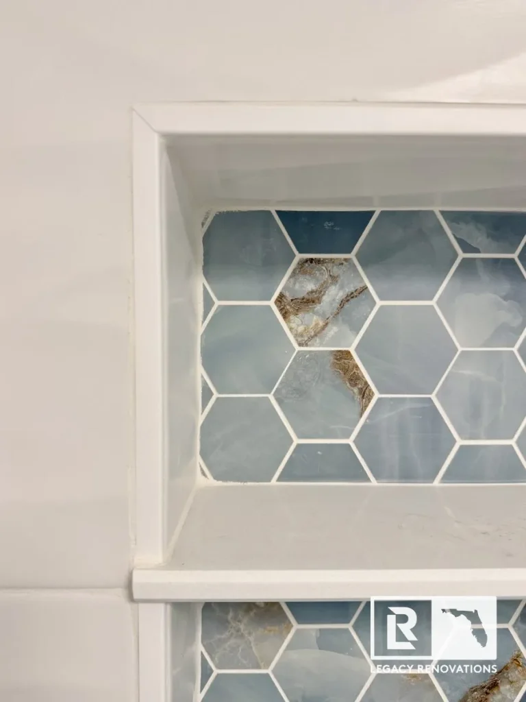 A detailed close-up of a recessed shower niche with white quartz shelving, featuring blue onyx-style hexagon mosaic backing tiles by Legacy Renovations in Jacksonville, FL.