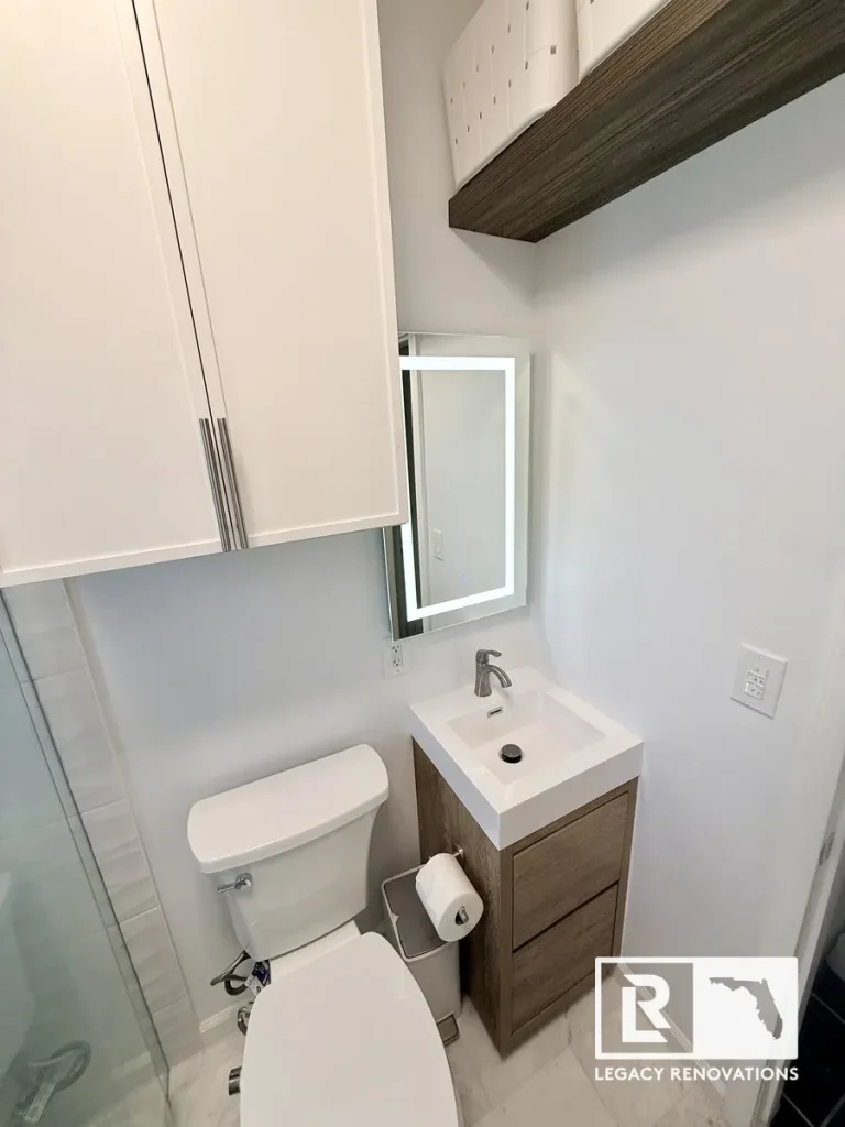 A finished small bathroom corner featuring a wood-grain vanity, white upper cabinet, floating shelf with storage baskets, and an LED-backlit mirror by Legacy Renovations in Jacksonville, FL.