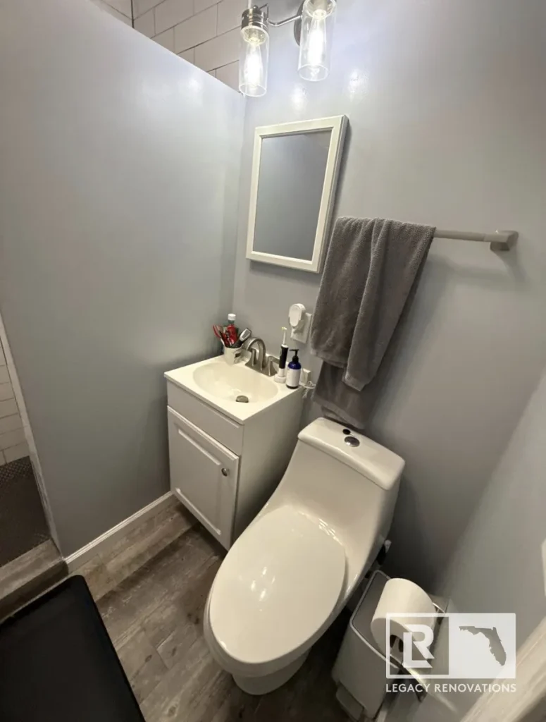 Completed small bathroom remodel with a white vanity, one-piece toilet, gray walls, wood-look flooring, and modern lighting in Jacksonville, FL.
