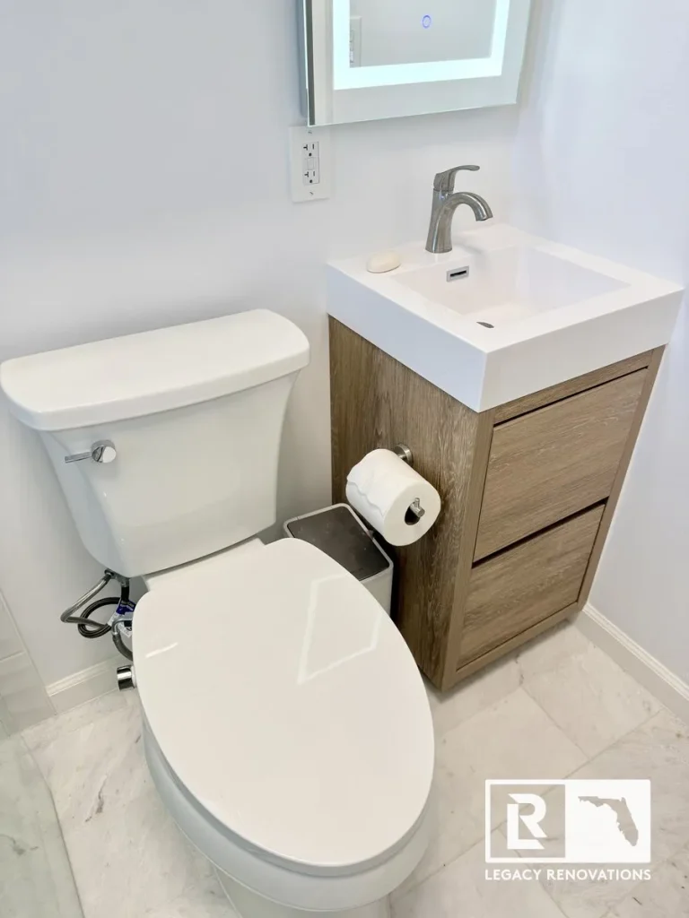 A completed small bathroom renovation featuring a white toilet, light wood grain vanity with integrated white sink, brushed nickel faucet, and marble-look floor tile by Legacy Renovations in Jacksonville, FL.