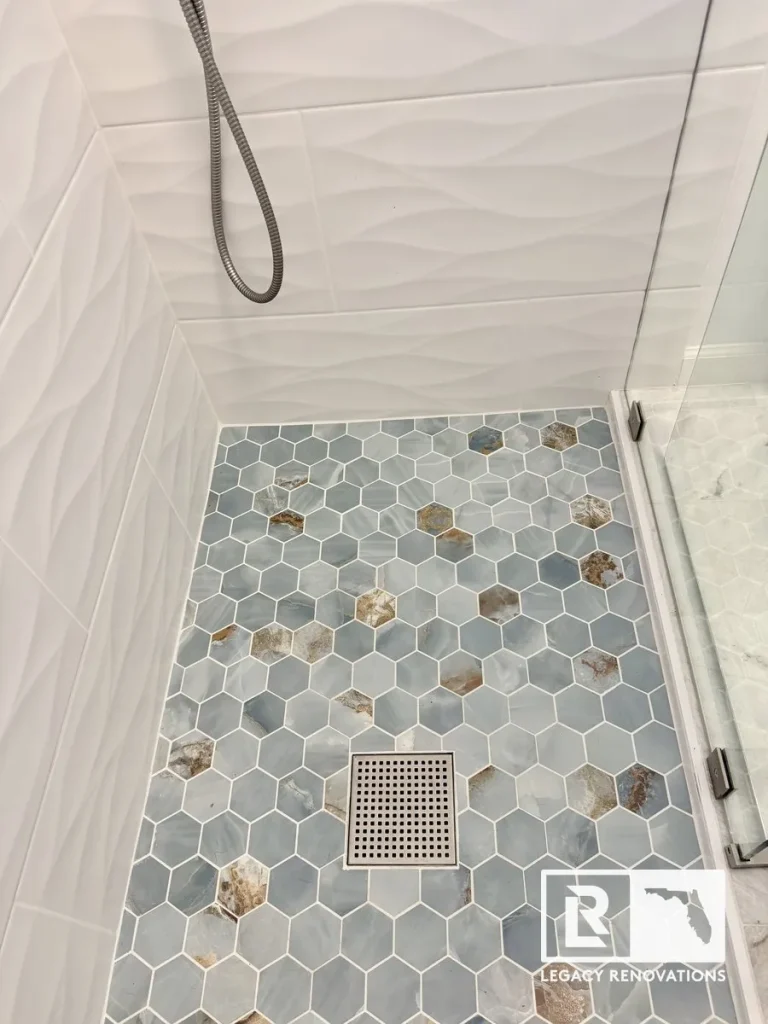 A finished walk-in shower floor featuring blue onyx-style hexagon mosaic tiles, a square stainless steel drain, and textured white wave wall tiles by Legacy Renovations in Jacksonville, FL.
