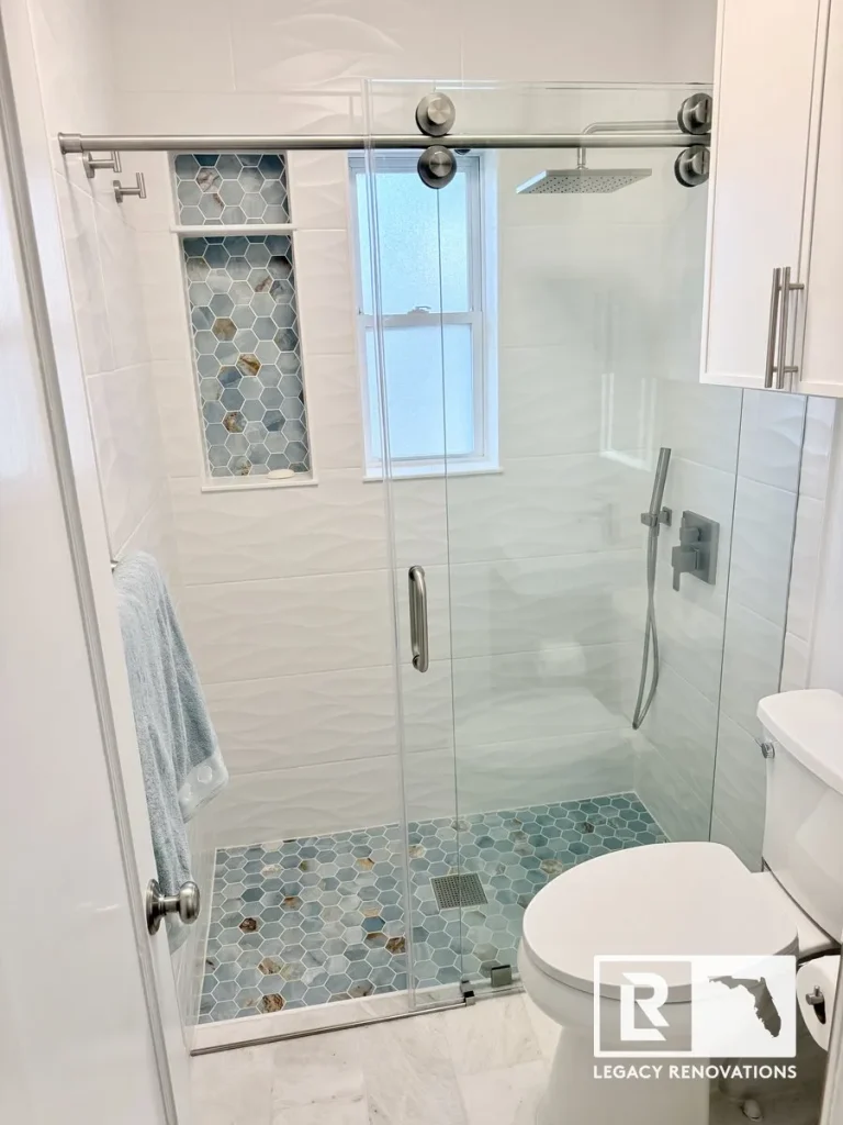 A completed bathroom renovation featuring a walk-in shower with a frameless sliding glass door, textured white wave wall tiles, blue onyx-style hexagon floor tiles, a double-shelf niche, and a brushed nickel rain shower head by Legacy Renovations in Jacksonville, FL.