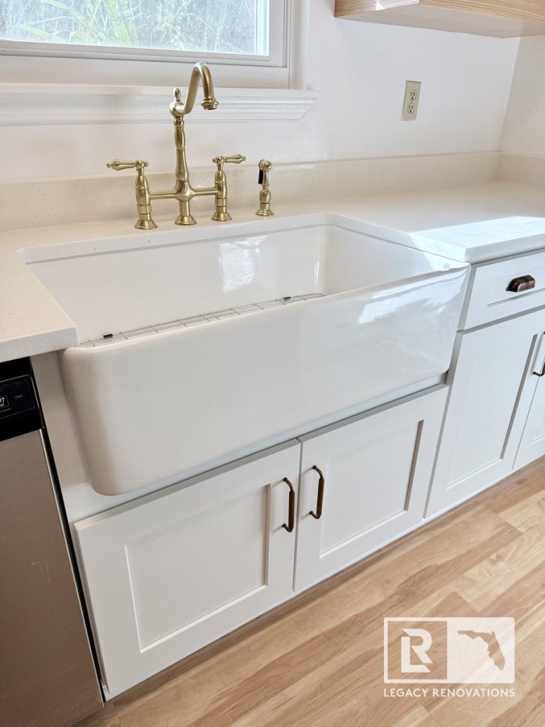 Deep white porcelain apron-front kitchen sink with a brass bridge faucet and white framed panel cupboards in a Jacksonville, FL home.