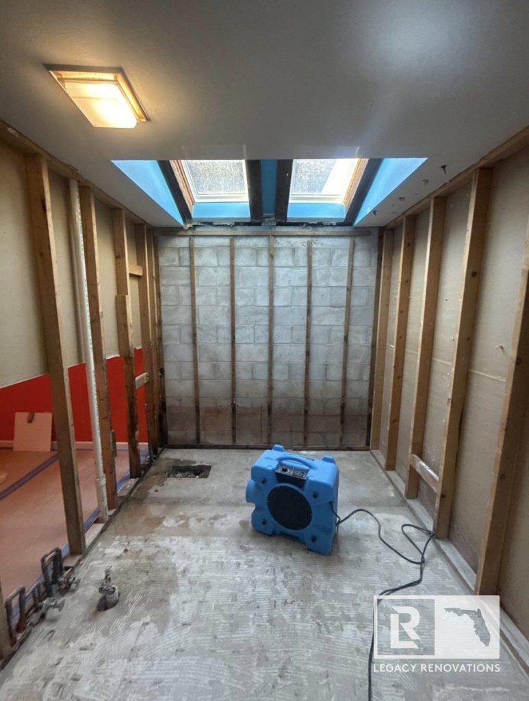 A residential bathroom during the gut renovation phase showing exposed wall studs, concrete block backing, a blue air mover fan, and dual skylights by Legacy Renovations in Jacksonville, FL.