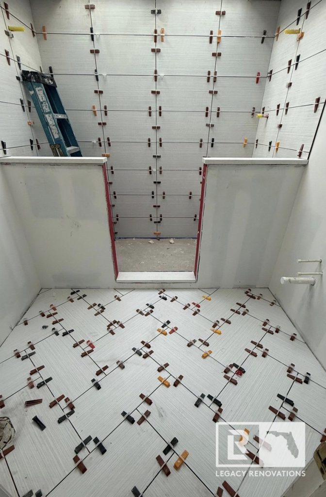 A bathroom renovation in progress featuring newly laid herringbone floor tile and textured wall tiles with plastic leveling spacers by Legacy Renovations in Jacksonville, FL.