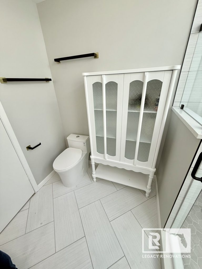 A white vintage linen cabinet with textured glass doors and a modern one-piece toilet on grey herringbone tile in a Jacksonville, FL bathroom renovation by Legacy Renovations.