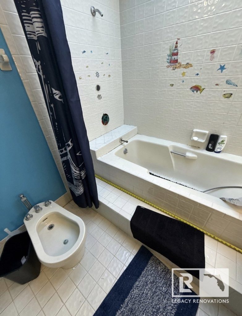 An outdated bathroom prior to renovation featuring a step-up tiled bathtub with a yellow measuring tape, a bidet, and sea-themed wall decals by Legacy Renovations in Jacksonville, FL.