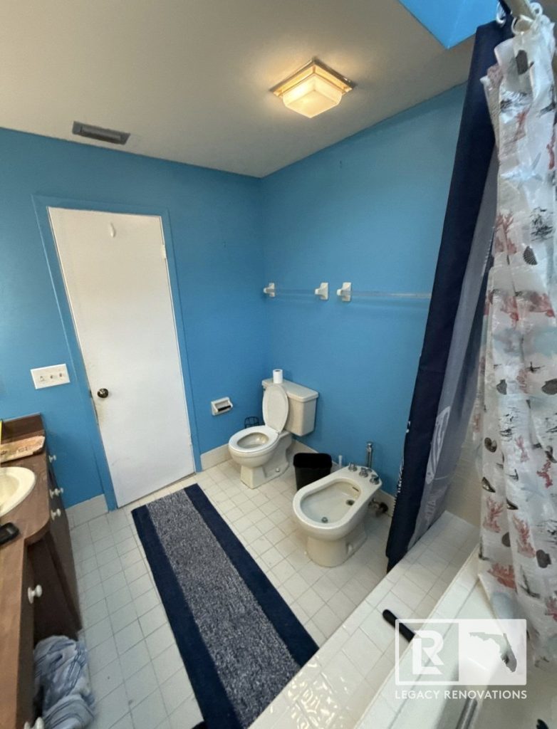 An outdated bathroom interior prior to remodel featuring bright blue walls, a toilet, an adjacent bidet, square floor tiles, and a wooden vanity by Legacy Renovations in Jacksonville, FL.