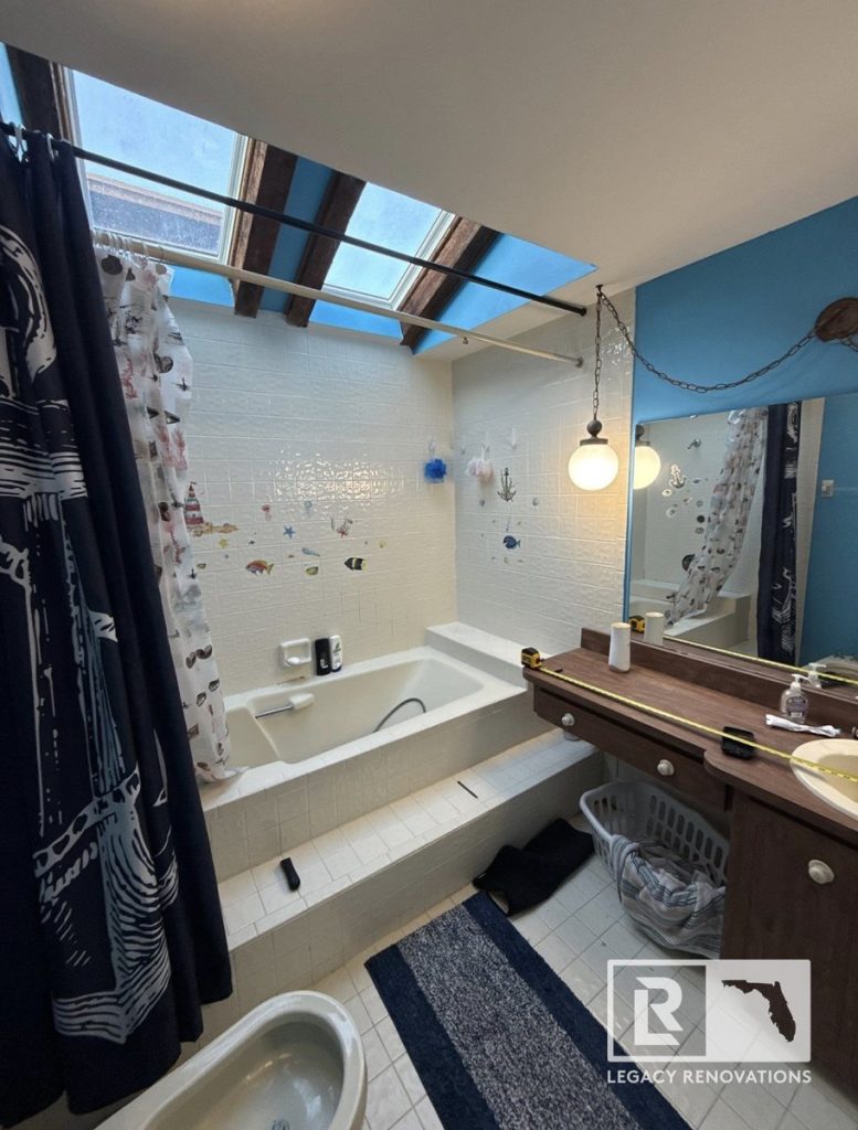 An outdated bathroom before remodeling, featuring a tiled step-up bathtub, wood vanity, hanging globe light, and ceiling skylights by Legacy Renovations in Jacksonville, FL.