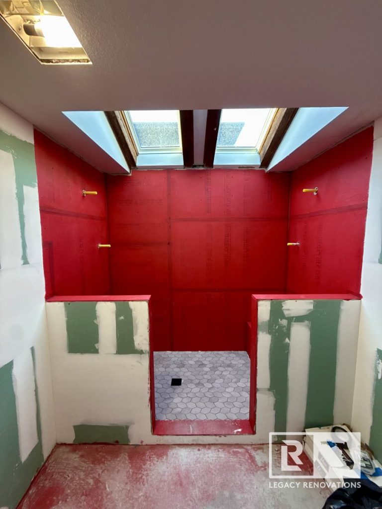 A walk-in shower under construction with bright red waterproofing on the walls, grey hexagon floor tiles, greenboard drywall framing, and twin skylights by Legacy Renovations in Jacksonville, FL.
