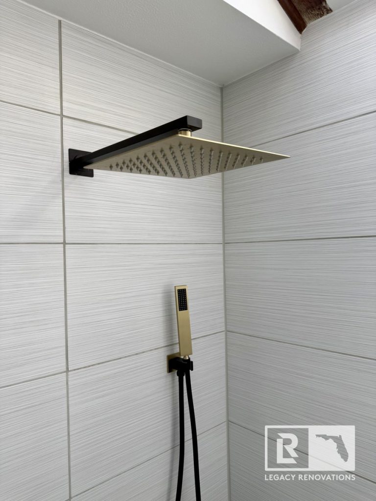 Close-up of a brushed gold and matte black square rainfall showerhead and handheld sprayer mounted on light grey textured wall tiles by Legacy Renovations in Jacksonville, FL.