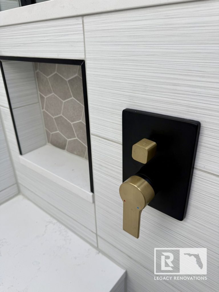 Close-up of a brushed gold and matte black shower lever next to a built-in storage niche with hexagon tile backing by Legacy Renovations in Jacksonville, FL.