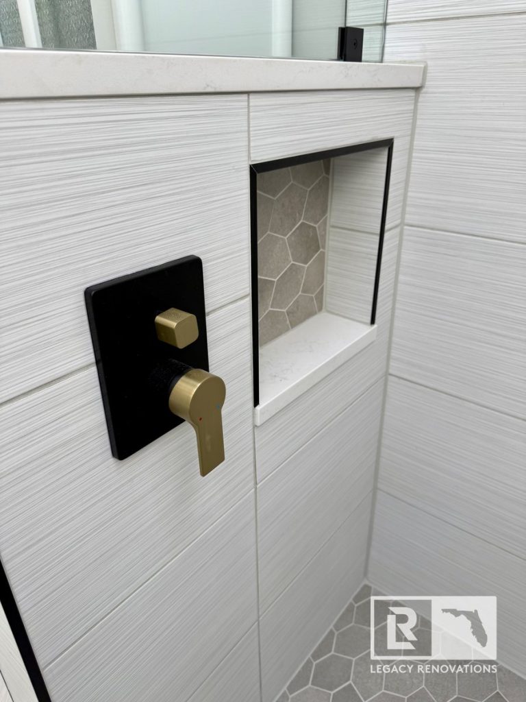 Close-up of a brushed gold and matte black shower control valve next to a recessed wall niche with hexagon tile inside a Jacksonville, FL bathroom renovation by Legacy Renovations.