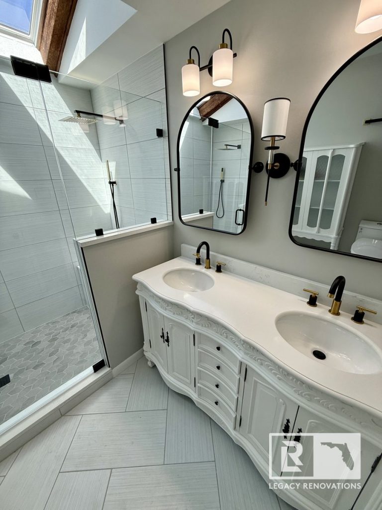 A full master bathroom renovation in Jacksonville, FL, featuring a carved white double vanity, arched black mirrors, and a glass-enclosed walk-in shower by Legacy Renovations.