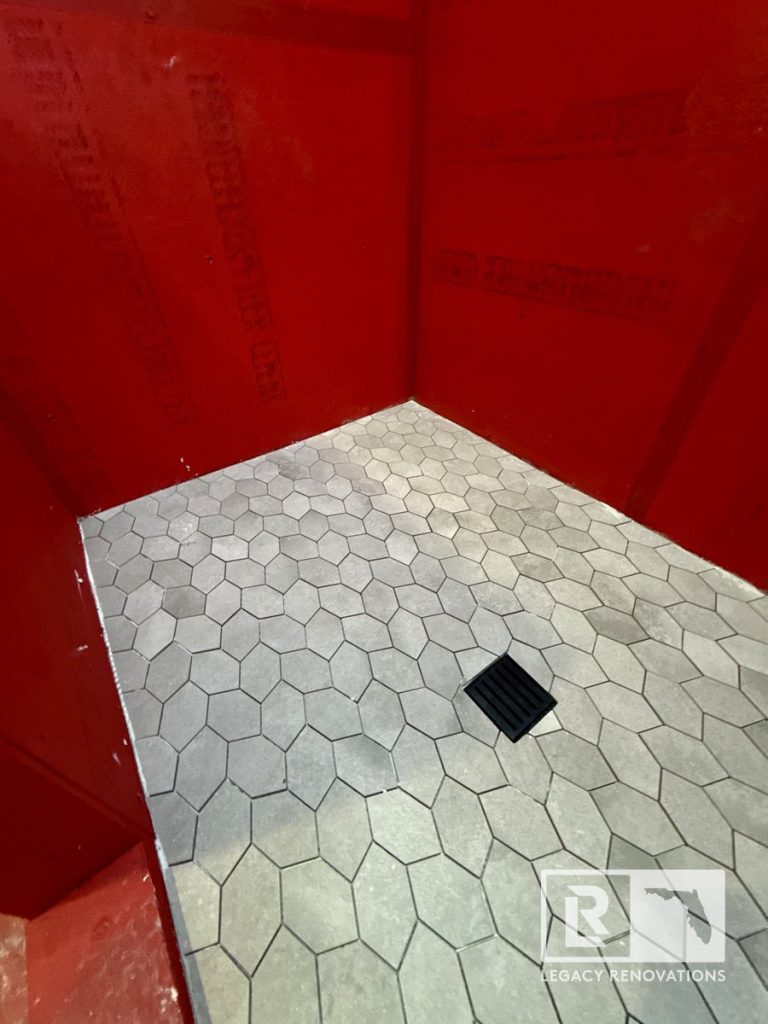 A close-up of a walk-in shower floor under construction featuring grey elongated hexagon tiles, a matte black square drain, and red waterproofed backing walls by Legacy Renovations in Jacksonville, FL.