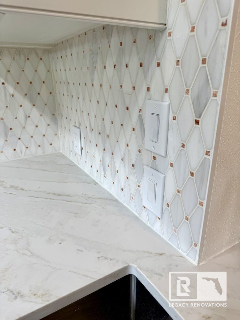 Close-up of a kitchen remodel in Jacksonville, FL featuring a geometric marble-look tile backsplash with metallic accents, quartz countertops, and a stainless steel farmhouse sink.